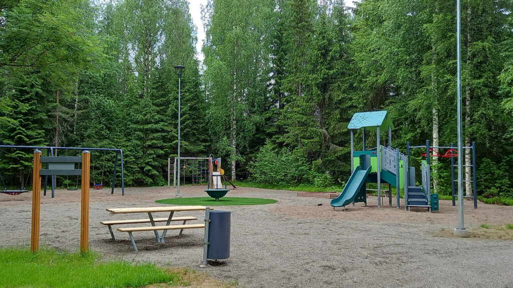 Metsän edessä leikkipaikka, jossa erilaisia leikkivälineitä.