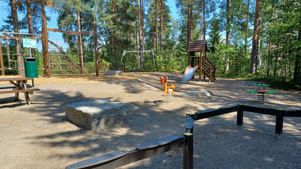 Leikkipuistossa olevia leikkivälineitä hiekkakentällä, taustalla metsää.