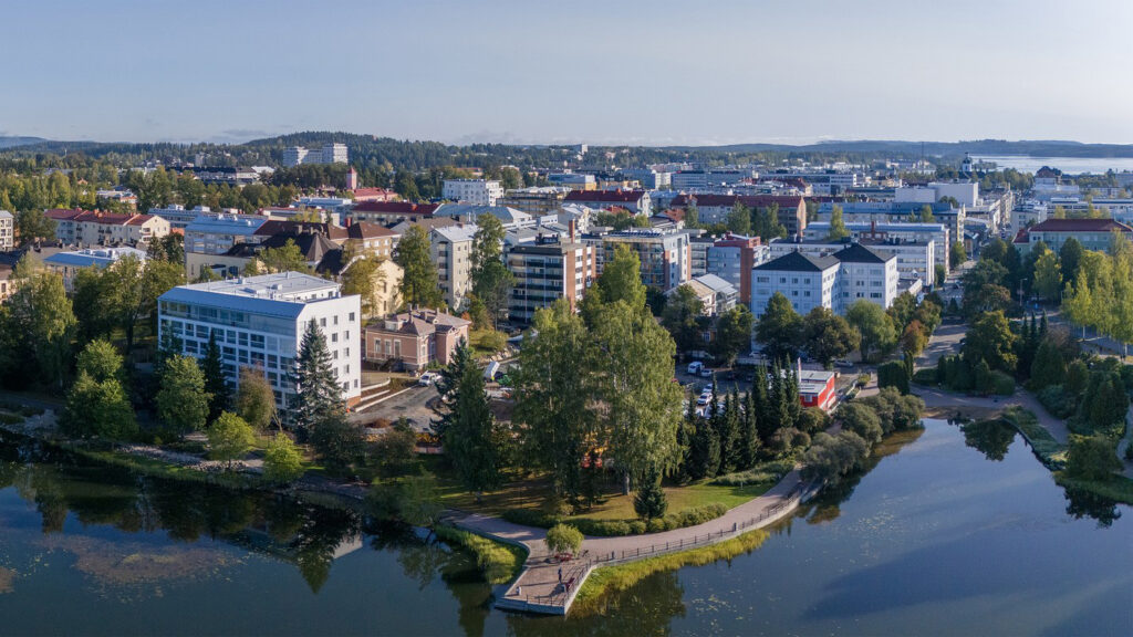 Kesäinen lammenranta aurinkoisena päivänä. Lammen rannalla vaaleita kerrostaloja.