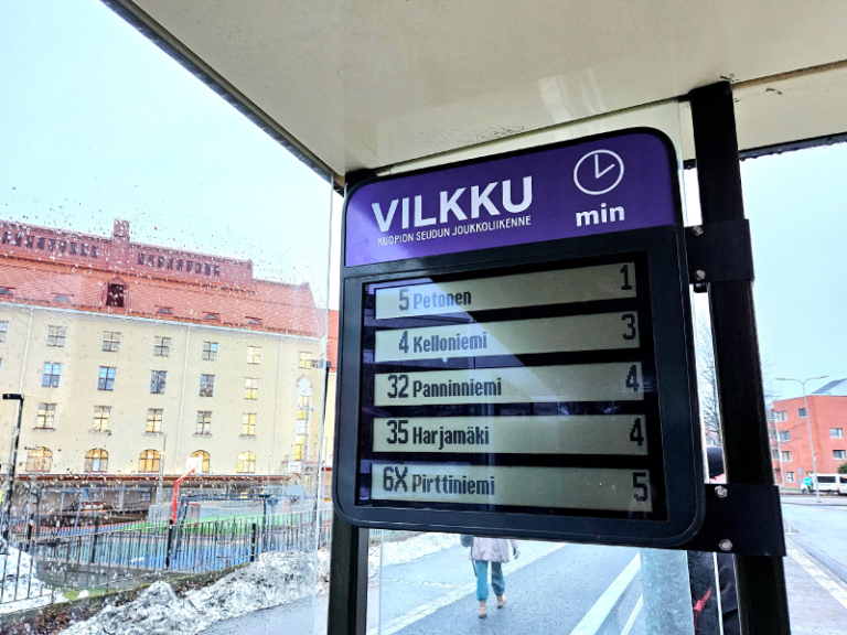 Bussipysäkin aikataulunäyttö, taustalla keltainen koulurakennus.