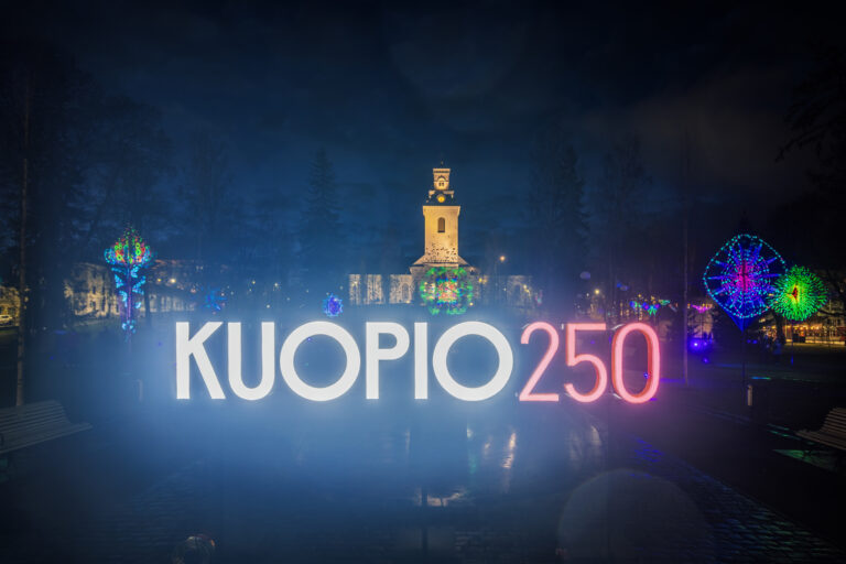 Valomainos, jossa teksti "Kuopio250". Taustalla tuomiokirkka ja valoteoksia.