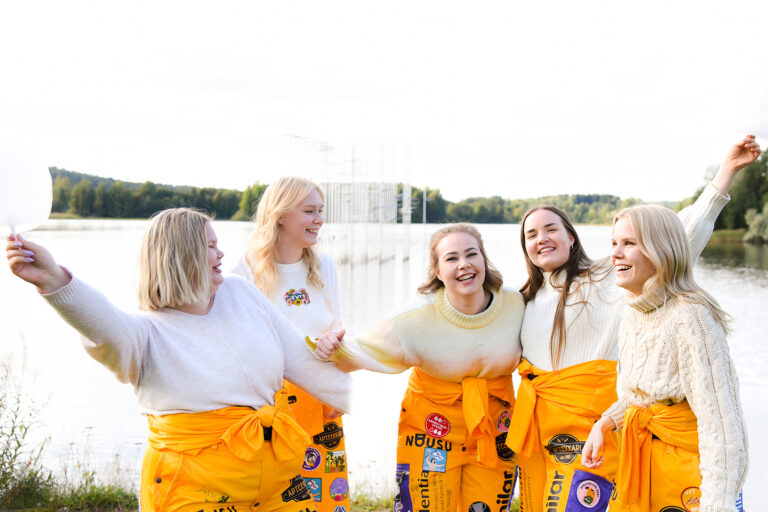 Viisi iloista, haalaripukuista opiskelijaa hassuttelee Savilahden rannalla.