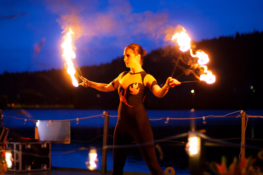 Nainen esiintyy tuliseipäiden kanssa pimenevässä illassa veden äärellä.