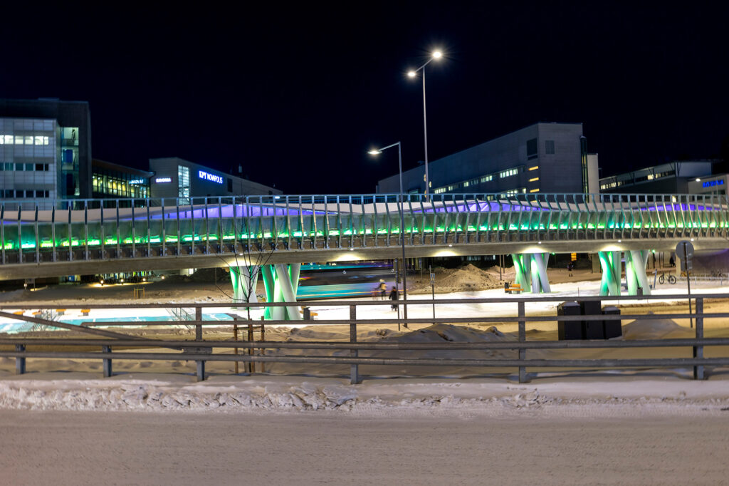 Silta loistaa sinivihreänä. Taustalla toimitilarakennuksia.