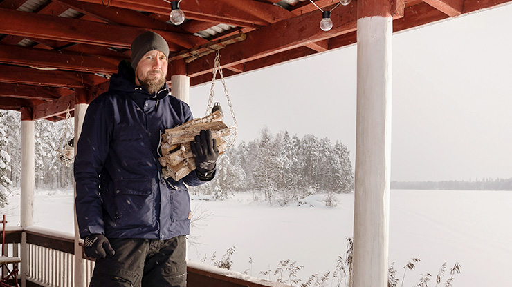 A man is standing on the porch with a stack of firewood in his hand, a winter lake in the background.