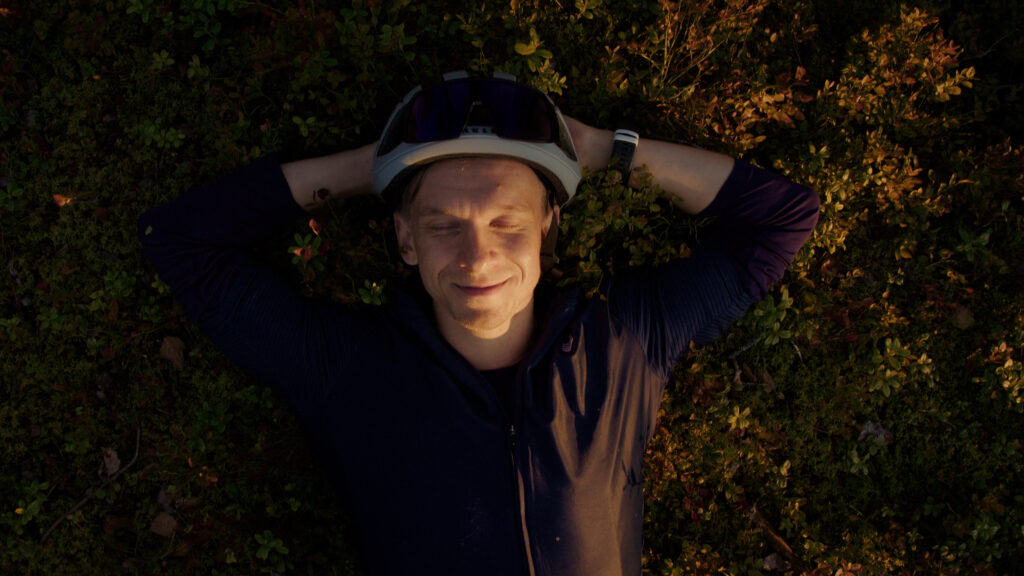 Aki Hurula is lying in the forest on a lingonberry mound, eyes closed and smiling.