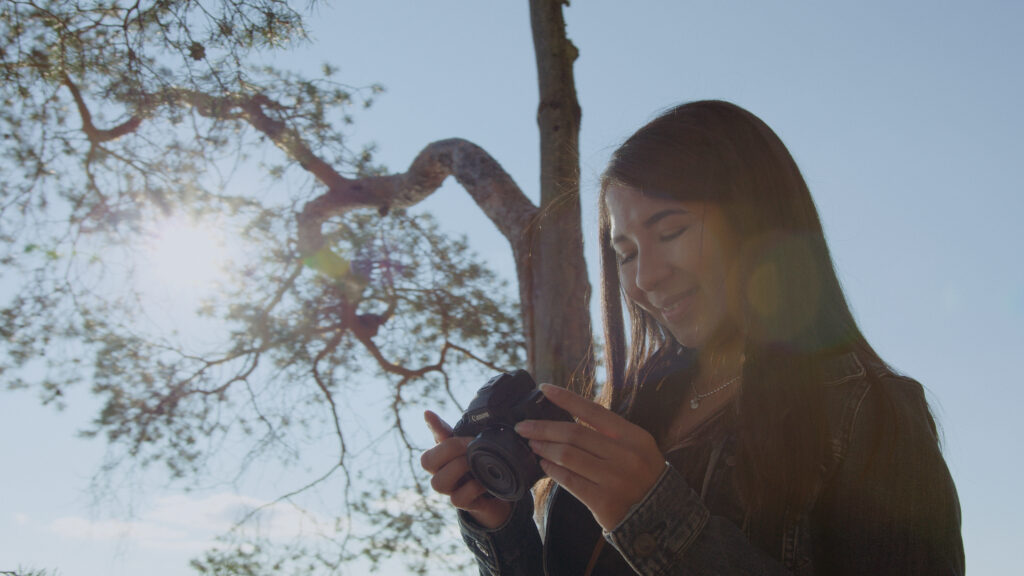 Jade Jiminez is looking at her camera, with the sun shining from behind a tree.