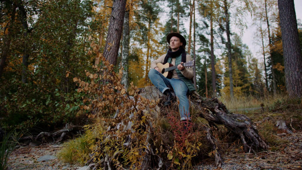 Roope Hakkarainen is sitting on a large stump, playing guitar in a pine forest.