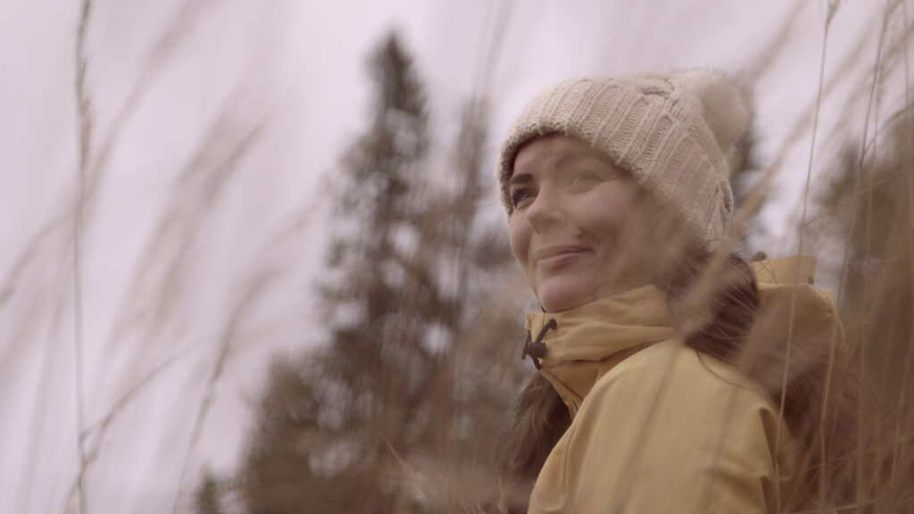 Satu Nisu is smiling while looking past the camera, with an autumnal atmosphere, trees and tall grasses in the background.